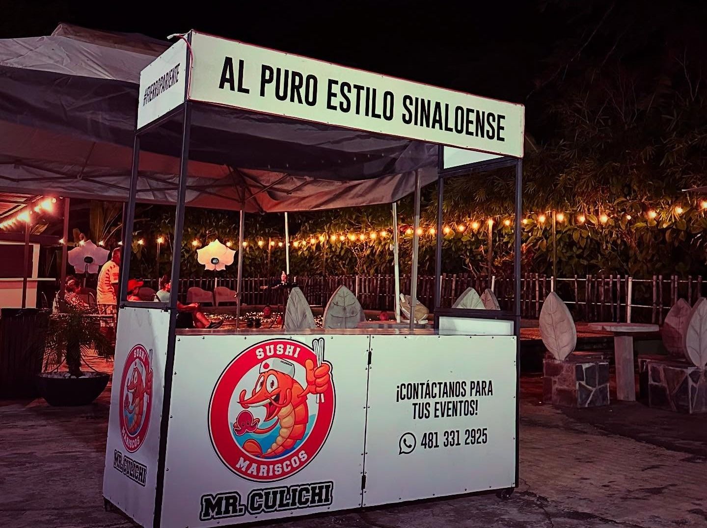 Carrito de Mr. Culichi de noche con el letrero Al puro estilo sinaloense y focos al fondo