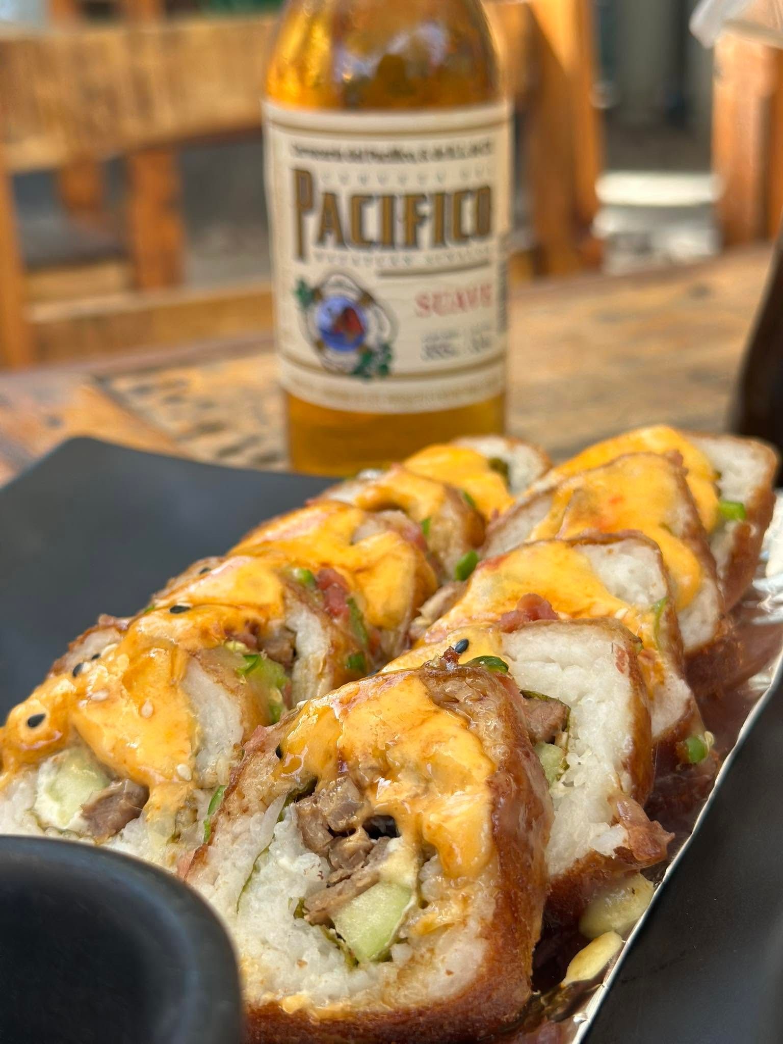 Rollo empanizado con salsa naranja sobre plato negro y una cerveza Pacífico al fondo